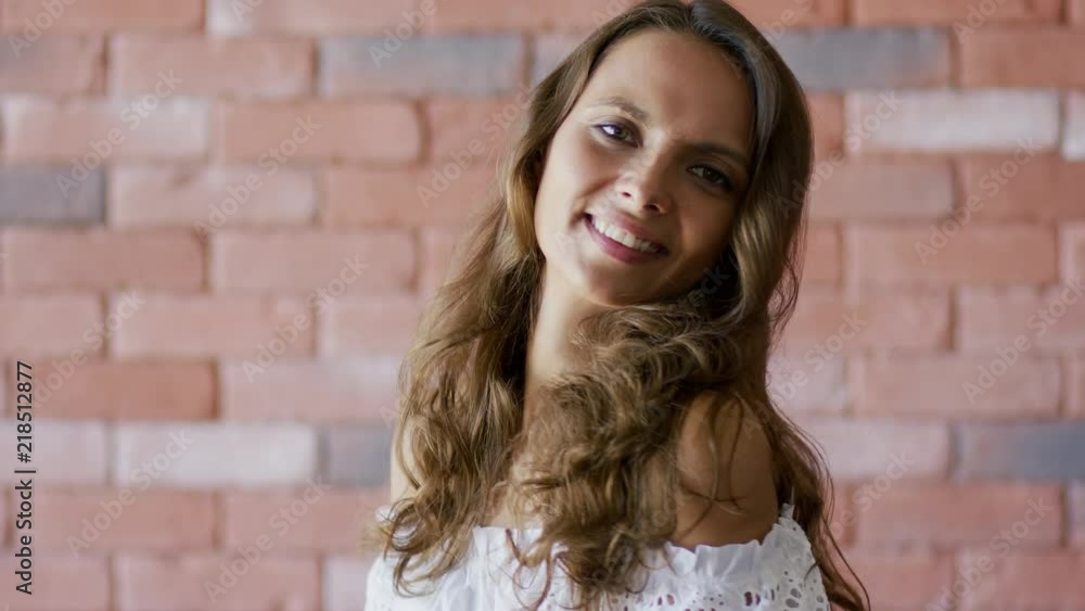 Pretty young woman with long wavy hair smiling and looking at camera while standing on background of brick wall Pretty young woman with long wavy hair smiling and looking at camera while standing on background of brick wall
