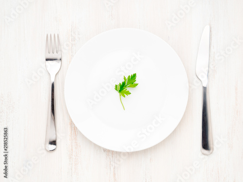 Parsley on empty white plate, fork and knife on a white wooden table, top view, diet concept