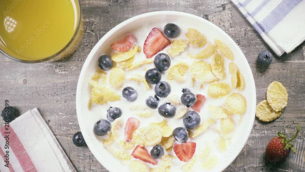 Slow motion blueberries fall into a bowl of cornflakes with milk and strawberries near a glass of orange juice top view Slow motion blueberries fall into a bowl of cornflakes with milk and strawberries near a glass of orange juice top view