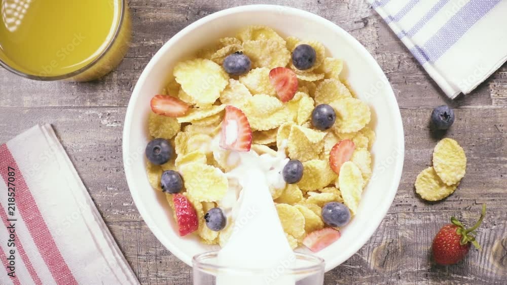 Slow motion in a bowl of cornflakes with strawberries and blueberries pour milk near a glass of fruit juice top view Slow motion in a bowl of cornflakes with strawberries and blueberries pour milk near a glass of fruit juice top view