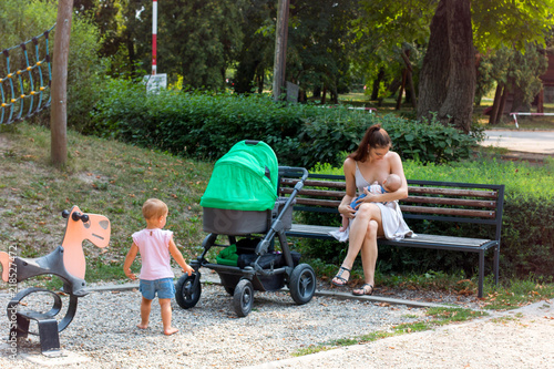 Motherhood, young pretty mother on parental leave breastfeeding her baby in children playground, curious baby is watching at her, mother and child, stop while walking with stroller on beautiful day
