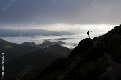 Wide mountain panorama. Small silhouette of tourist with backpack on rocky mountain slope with raised hands over valley covered with white puffy clouds. Beauty of nature, tourism and traveling concept