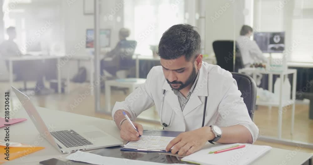Young physician therapist sitting at table, studying his work schedule for next month, writing in documents and typing in laptop. Busy doctor Young physician therapist sitting at table, studying his work schedule for next month, writing in documents and typing in laptop. Busy doctor