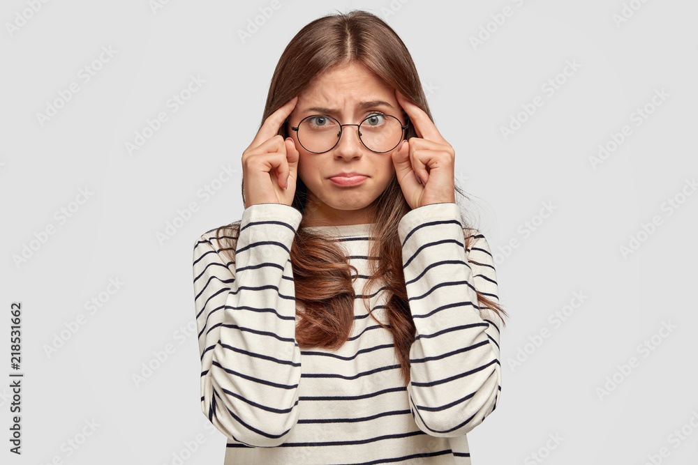 Thoughtful young woman with bad memory, keeps fingers on temples, has dissatisfied expression, dressed in striped casual sweater, isolated over white background. People and thoughts concept. Thoughtful young woman with bad memory, keeps fingers on temples, has dissatisfied expression, dressed in striped casual sweater, isolated over white background. People and thoughts concept.