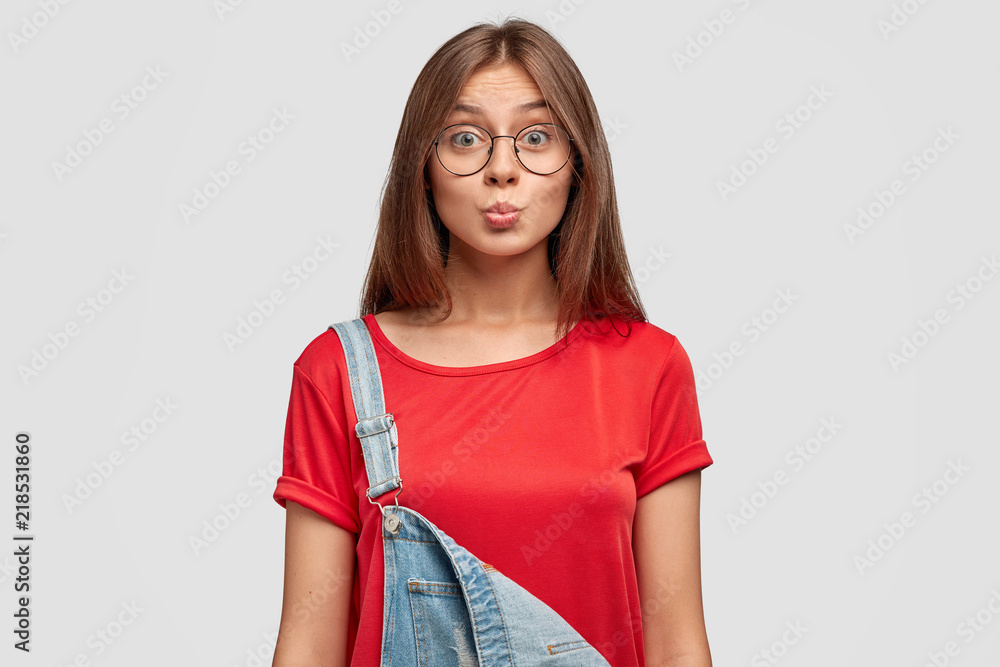 Φωτογραφία Lovely cute young female with friendly expression, makes grimace, pouts lips, dressed in stylish red t shirt and overalls, poses against white background