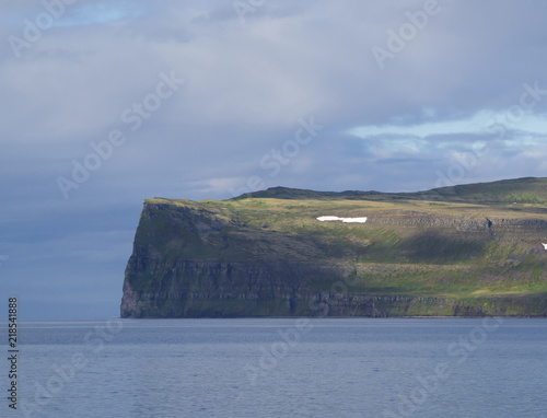 Scenic view from ocean on beautiful snow patched cliff in west fjords, nature reserve Hornstrandir in Iceland, blue sea and cloudy sky background, golden hour light photo