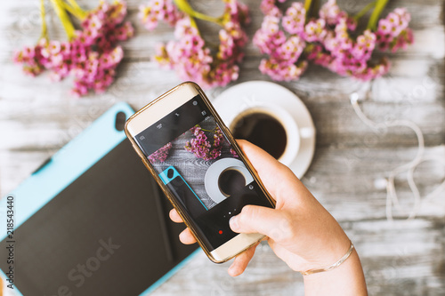 Instagram photographer blogging workshop concept, hand holding phone and taking photo of stylish flowers, cup of coffee and graphic tablet on grey wooden rustic background.space for text. photo