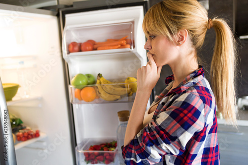 Pretty young woman hesitant to eat in front of the fridge in the kitchen.
