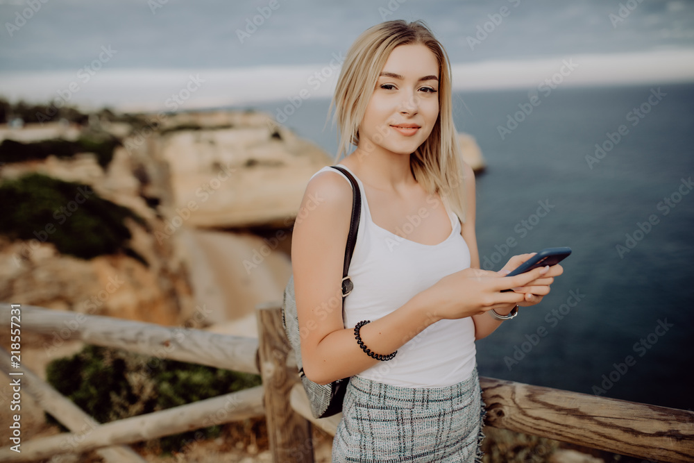 Young woman updating her social network status while relaxing at the rock beach with panorama view. Travel concept.