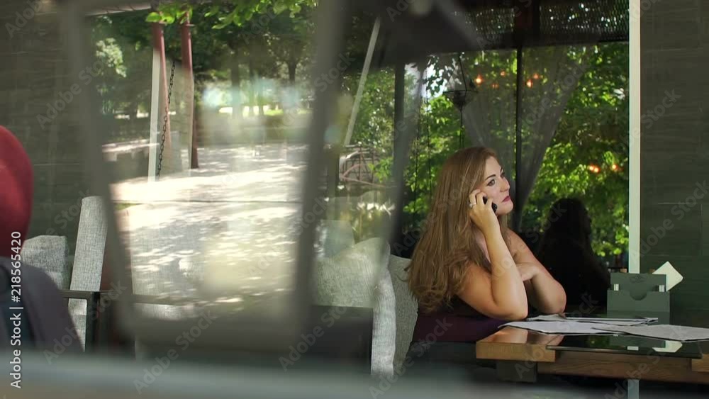 Young overweight girl in t-shirt and jeans sitting in a street cafe and talking on the phone, slow motion. Girl XXL. Young overweight girl in t-shirt and jeans sitting in a street cafe and talking on the phone, slow motion. Girl XXL.