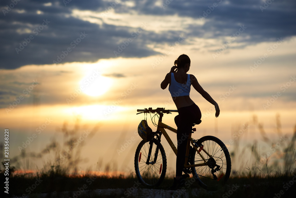 Obraz premium Silhouette of sporty female cyclist, wearing sportswear and posing near her bicycle on trail. Slender incognito woman enjoying nature and observing wonderful landscapes and amazing sunset.