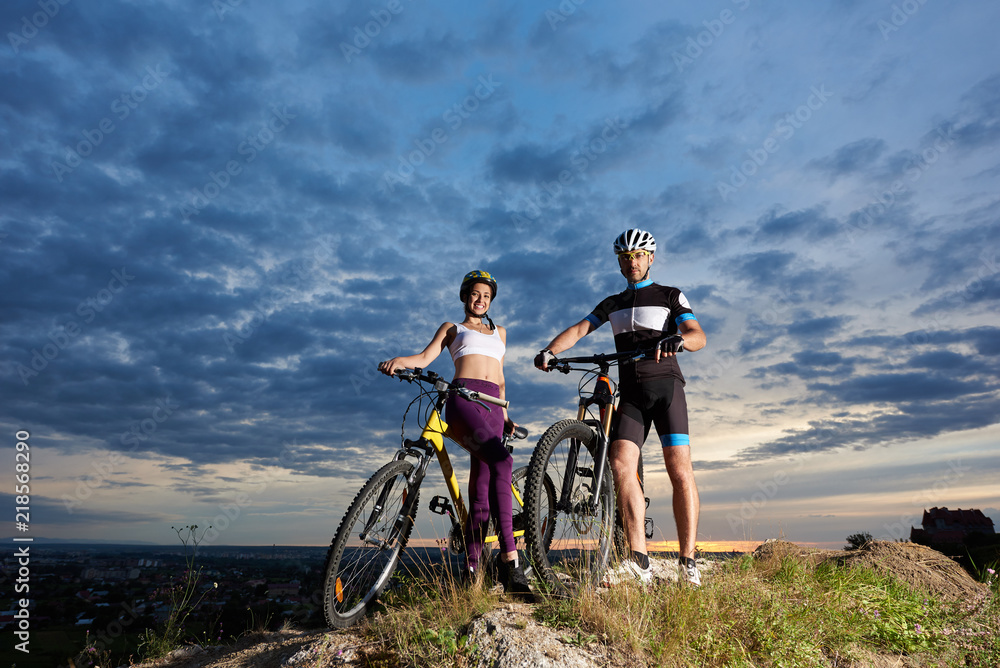 Two positive cyclists holding hands on handle bars of their bikes, looking at camera and smiling. Beautiful girl wearing violet leggings and white top, man in black sportswear and sport glasses.