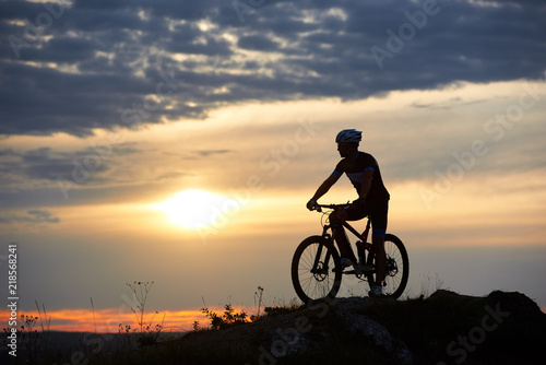 Robust and strong man wearing sportswear and helmet sitting on his bike and posing on hill. Cyclist enjoying great view of sunset. Concept of motivation, recreation and healthy activities.