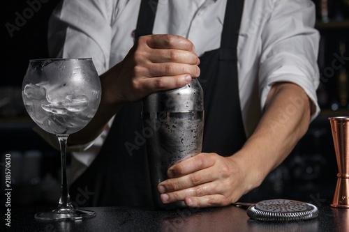 The bartender closes the cooled shaker near is a wine glass filled with ice, a jiger and a strainer on a bar counter.