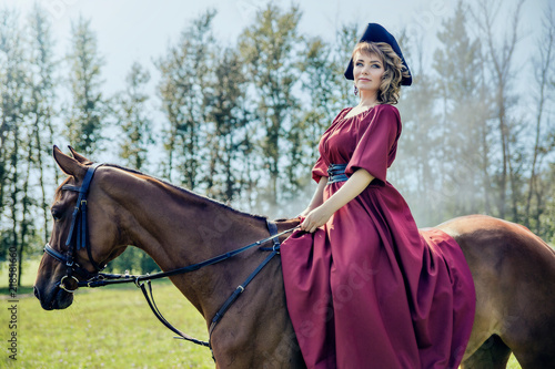 Beautiful girl in a red long red dress and in a black hat with a cocked hat riding a brown horse. Looks at the camera.