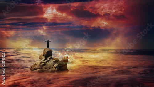 A man with spreading arms standing on a rock protruding above the clouds and looking into the distance.