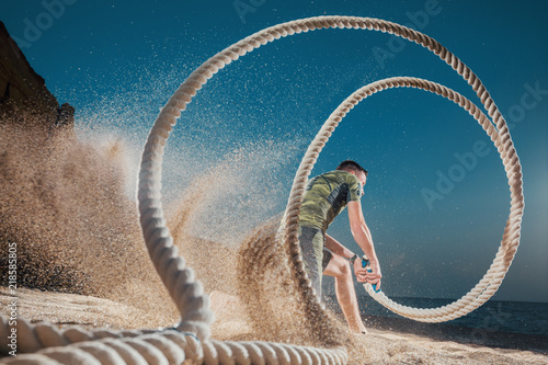 Man training with battle rope on the beach. Athlete doing cross fitness workout outdoor. Sport fit exercise.