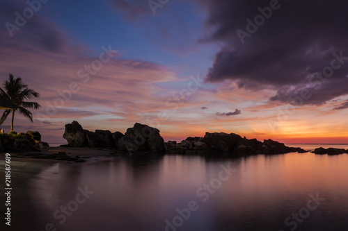 Vibrant and colorful sunset sky is reflected in the smooth water of the sea shot on the long exposure with the silhouette of the rocks and a palm tree