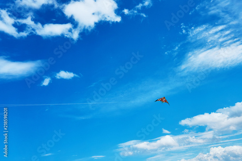 A large kite floats high in the sky among the clouds against the blue sky and the bright summer sun and it is controlled by two white ropes going to the ground