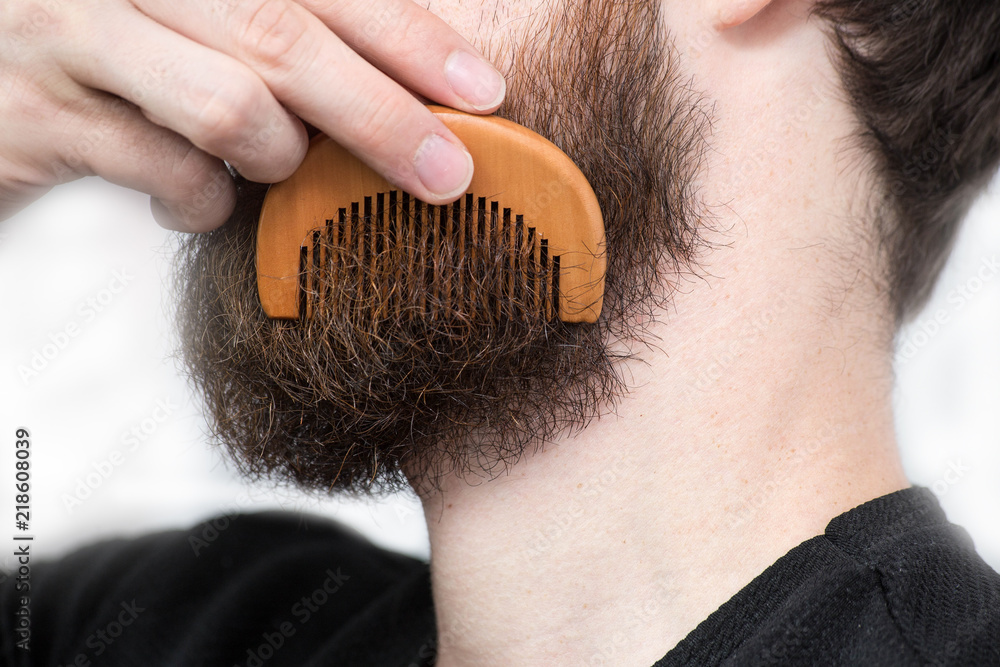 Closeup of a young man styling his long beard with a comb while standing alone in a studio against a white background - obrazy, fototapety, plakaty 