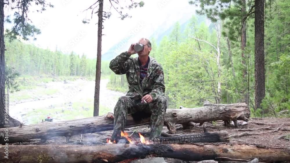 A man drinks alcohol in a forest by the fire. The tourist drank strong alcohol and poured it into the fire from the mug. A man drinks alcohol in a forest by the fire. The tourist drank strong alcohol and poured it into the fire from the mug.