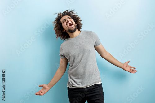 A curly-headed handsome man wearing a gray T-shirt is standing and laughing hard with his hands widly spread, looking upwards over the blue background. Happiness and joy.
