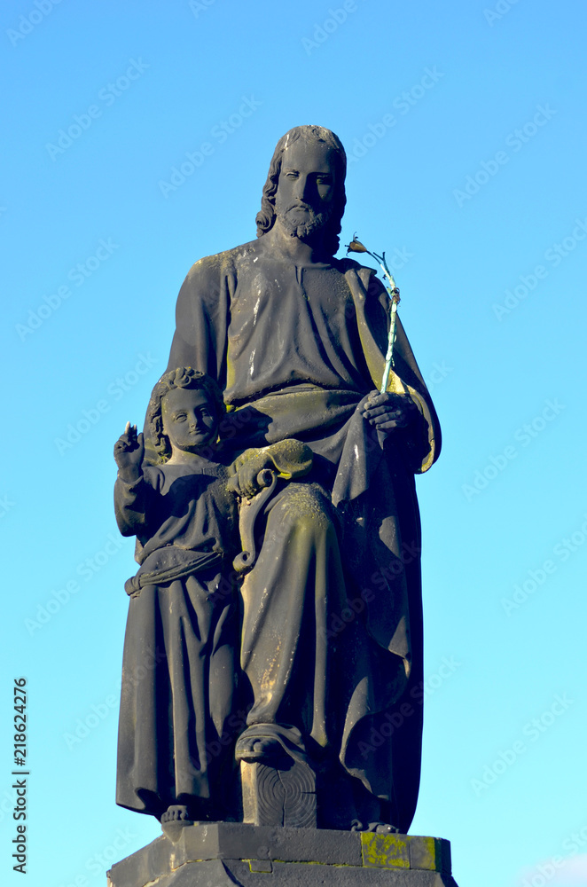 Fototapeta premium A weathered statue of St. Jospeh and Jesus is on the Charles Bridge, Prague. St Jospeh hold a flowering rod in one hand. Behind is a bright blue sky.