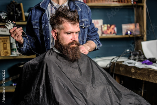 Barber preparing hair clipper for bearded man, barbershop background. Barber with clipper and brutal bearded client. Hipster lifestyle concept. Hipster client covered with cape getting haircut