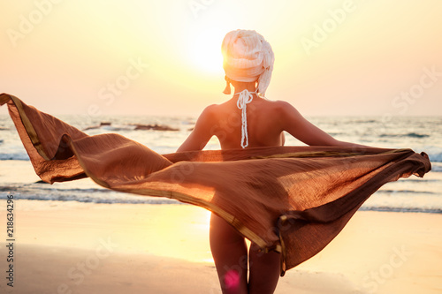 sexy and beautiful young woman in a turban and swimsuit with luxury shawl pareo chiffon dress develops in the wind . awesome sunset on the sea freedom by the beach photo