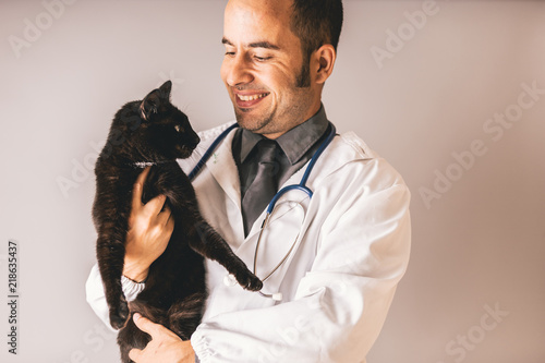 A veterinary doctor with a stethoscope around his neck holds a black cat and smiles