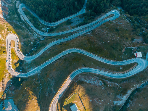 Aerial: top down view tailspin over winding mountain road on the Italian Alps, crossing green meadow and pasture in alpine valley, adventure road trip in summer.