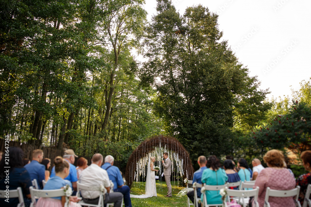Obraz premium Wedding. Rustic wedding. Bride and groom near the wedding arch at the wedding ceremony, guests on chairs looking on newlyweds couple