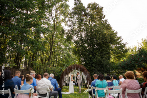 Wedding. Rustic wedding. Bride and groom near the wedding arch at the wedding ceremony, guests on chairs looking on newlyweds couple