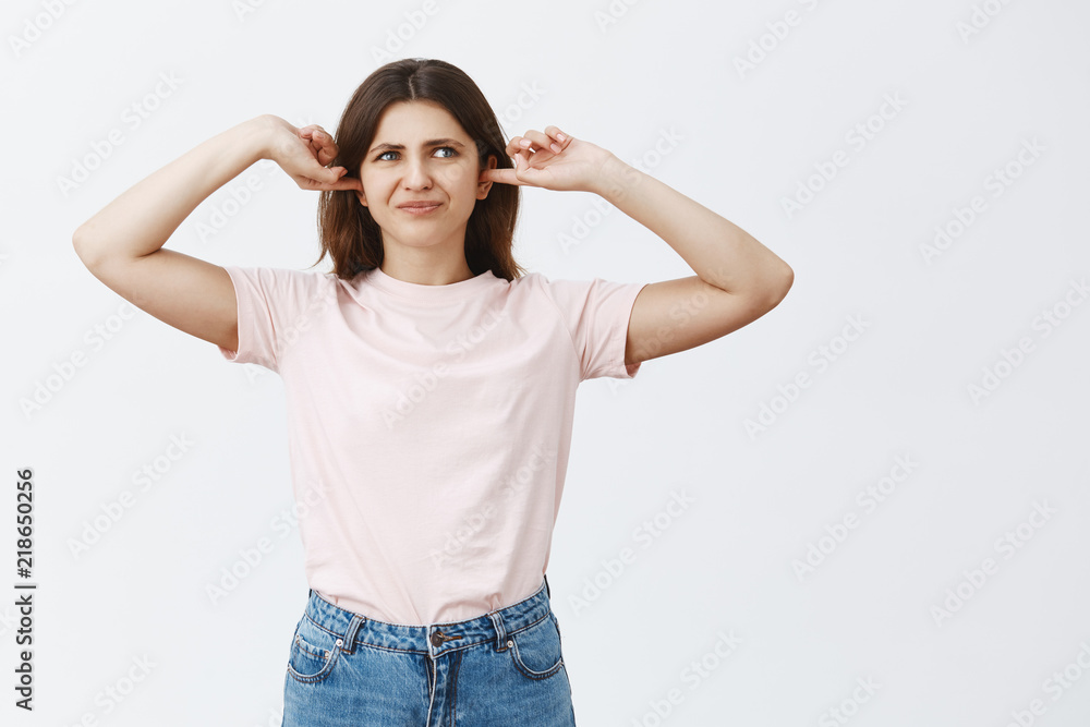 Woman being disturbed cannot concentrate hearing loud terrible noise covering ears with index fingers purisng lips and frowning from dislike and discomfort standing over gray background Woman being disturbed cannot concentrate hearing loud terrible noise covering ears with index fingers purisng lips and frowning from dislike and discomfort standing over gray background
