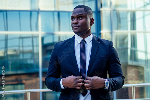 portrait of beautiful and stylish African American man in a fashionable black jacket and a white shirt with a collar with a elegant tie posing background of Manhattan glass offices cityscape