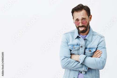 Your cocktails suck. Displeased good-looking stylish male in trendy sunglasses and denim jacket, sticking out tongue and frowning from dislike and displeasure, holding hands crossed in offended pose