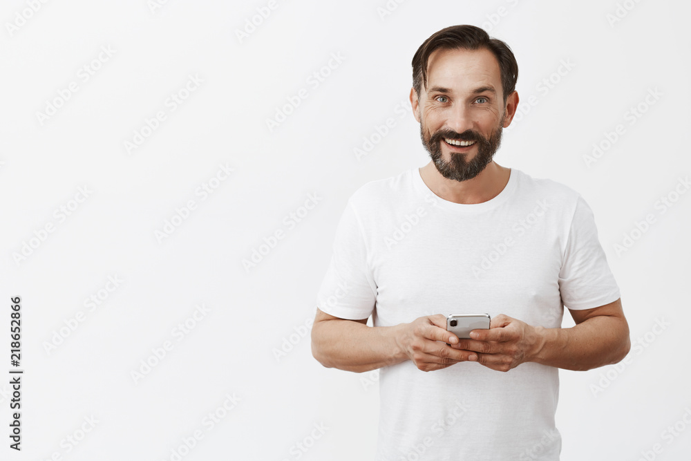 Arrenging date via messages, so thrilling. Portrait of charming friendly-looking adult male with stylish haircut and beard, holding smartphone and gazing with broad interested smile at camera Arrenging date via messages, so thrilling. Portrait of charming friendly-looking adult male with stylish haircut and beard, holding smartphone and gazing with broad interested smile at camera