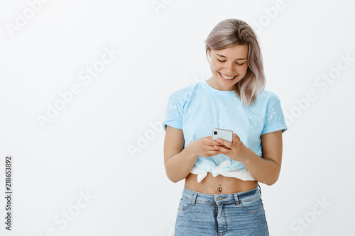 Good-looking carefree european female with blond hair and tattoos, holding smartphone and typing message, smiling broadly while looking at screen, being pleased and happy receive news form friend