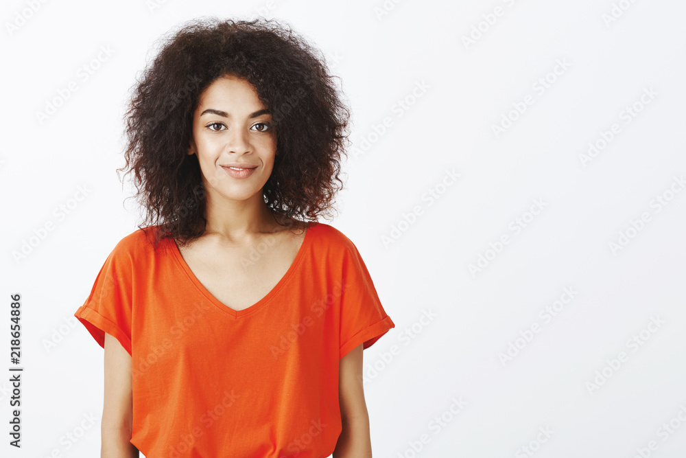 Fototapeta premium Beautiful people and positive emotions concept. Joyful good-looking dark-skinned model with afro hairstyle standing in stylish t-shirt and smiling happily at camera, picking puppy in local shelter