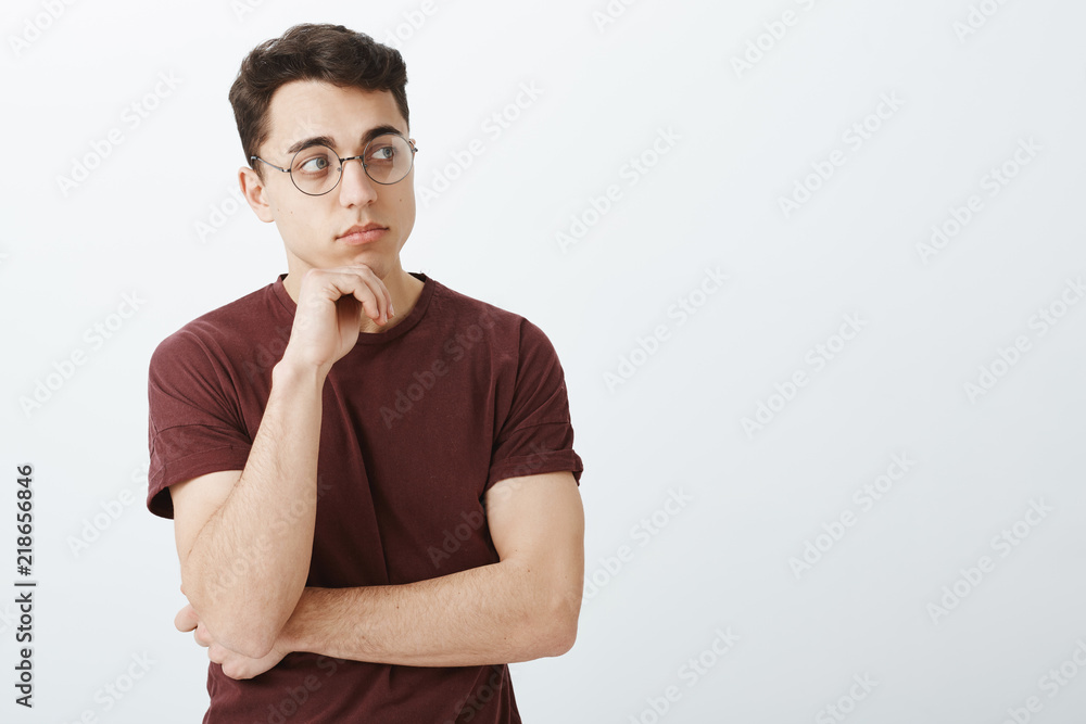Portrait of smart creative male entrepreneur in t-shirt and round glasses, looking in right direction and thinking with half-crossed hands, holding palm on chin while making decision or solving issue Portrait of smart creative male entrepreneur in t-shirt and round glasses, looking in right direction and thinking with half-crossed hands, holding palm on chin while making decision or solving issue