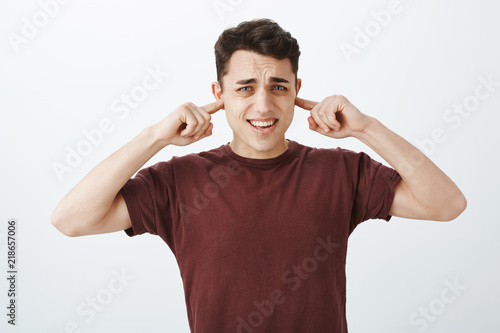 Turn that annoying sound off. Bothered displeased caucasian male coworker in red t-shirt, covering ears with index fingers, frowning and clenching teeth from awful noise, standing over gray wall