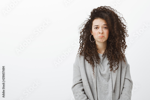 Girl feels regret missing interesting movie in cinema. Sulking sad attractive curly-haired female in stylish clothes and earrings, grimacing, feeling upset and disappointed, whining over gray wall photo