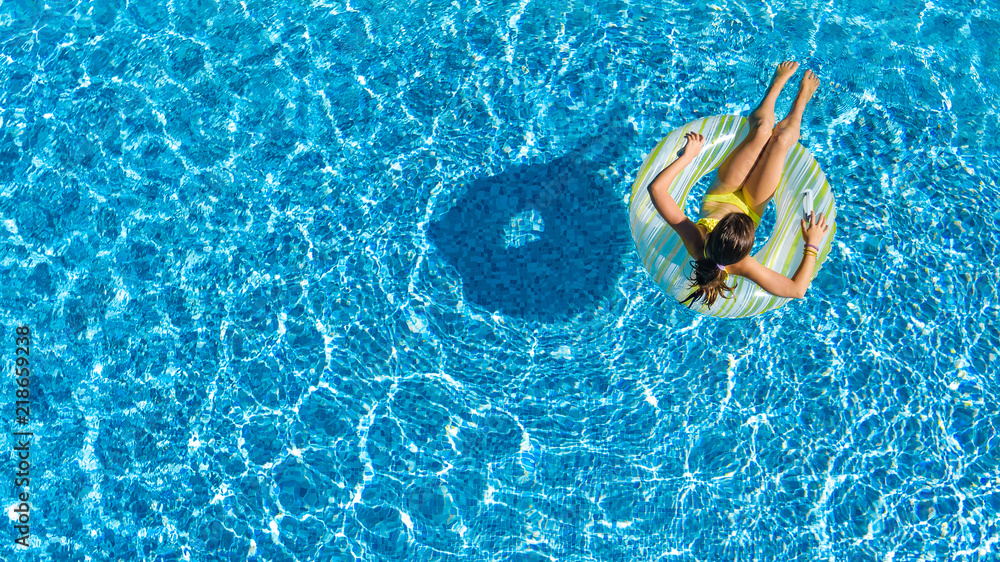 Aerial top view of little girl in swimming pool from above, kid swims on inflatable ring donut , child has fun in blue water on family vacation resort
