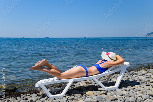 A beautiful girl in a blue bikini and a white hat is sunbathing on the beach, lying on a white chaise longue. The blonde lies with her back to the top. The woman lies on her stomach.