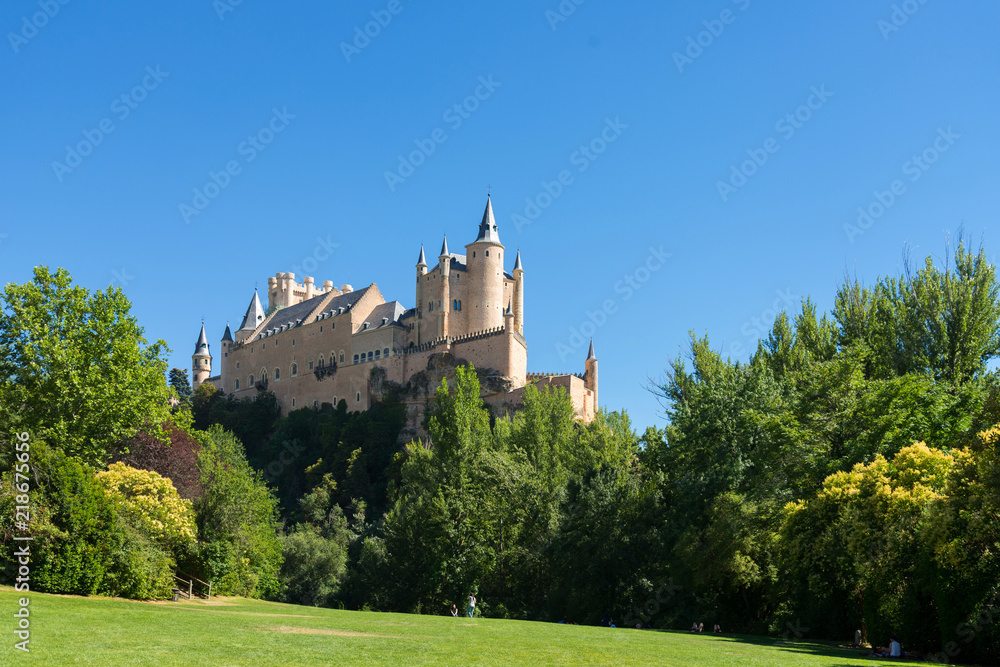 Scenery of Alcázar de Segovia on the hill from Park de San Marcos, during summer season with blue clear sky in Segovia, Spain. The castle on the hill located behind range of tree and park in Segovia.