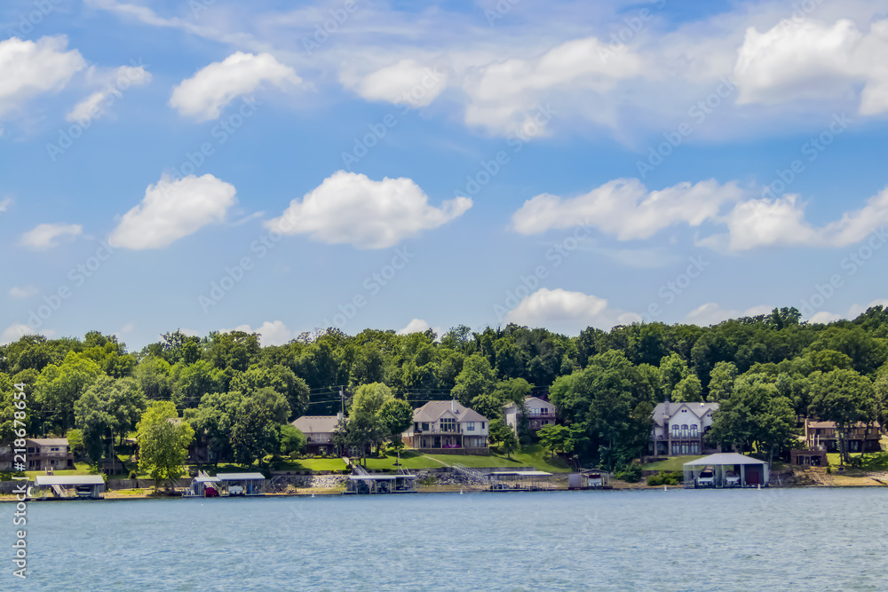 Upscale homes with boat docks built along the edge of a lake with tall green trees under a blue sky with fluffy clouds