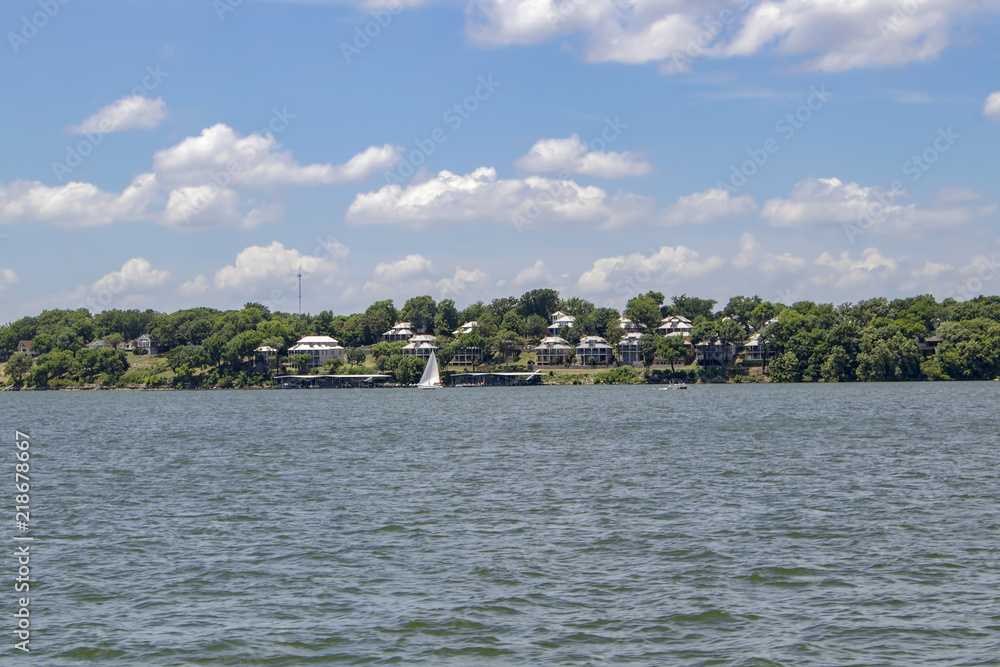 Condos beside the lake among the trees under a cloudy blule sky with a sailboat and a pontoon out on the water Condos beside the lake among the trees under a cloudy blule sky with a sailboat and a pontoon out on the water