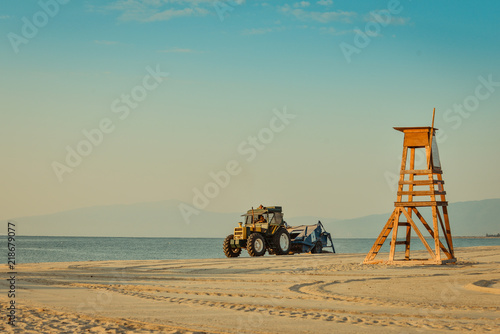 Fototapeta Naklejka Na Ścianę i Meble -  Tractor cleaning the beach on a calm morning at the beach with a safeguard post on a sunny morning with Olympus mountain and clear sky in the background.