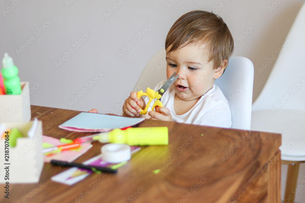 Little baby boy holding scissors in his hands and making handmade paper greeting card for mother. Fine motor skills development. DIY concept. Child safety. Little baby boy holding scissors in his hands and making handmade paper greeting card for mother. Fine motor skills development. DIY concept. Child safety.