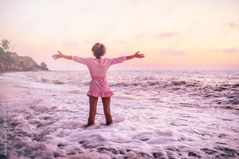 Silhouette of a girl in a pink dress on the beach, on the background of a pink dawn, with arms spread to the sides and knee-deep in a foaming wave.